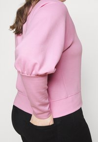 Jasnoróżowa bluza z balonowymi rękawami, ściągaczami przy mankietach i dopasowanym dołem, połączona z czarnymi spodniami. Miękka faktura i casualowy styl.