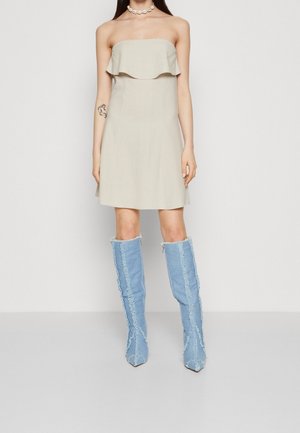 Femme portant une robe beige sans bretelles avec un détail à volants et de grandes bottes en denim bleu clair aux coutures effilochées, debout devant un fond uni.