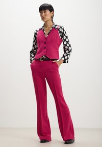 Gilet fucsia con bottoni neri sopra una camicia a pois. Pantaloni rosa svasati e cintura nera. Tessuto liscio con motivi a contrasto.