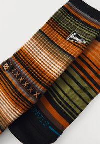 Gestreifte Socken in den Farben Orange, Grün und Schwarz. Sie weisen mehrere Muster auf, darunter horizontale und vertikale Linien, sowie ein Logo-Detail.