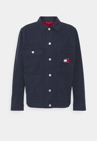Mörkblå denimjacka med knäppning fram, två bröstfickor och en logotypapplikation på vänster ficka. Har silverfärgade tryckknappar och en krage.