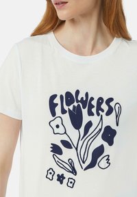 Biała bawełniana koszulka z granatowym grafiką stylizowanych kwiatów i napisem "KWIATY". Projekt zawiera różne kształty kwiatów i detale.