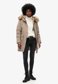 Piumino beige con cappuccio in pelliccia staccabile, dotato di due tasche frontali e chiusura a zip, indossato sopra un maglione bianco a trecce e jeans neri.