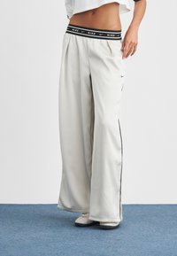 Λευκό παντελόνι φόρμας Nike με μαύρη μέση με λογότυπο, φαρδιά πόδια και ρίγες στο πλάι. Το ύφασμα φαίνεται λείο με ελαφριά γυαλάδα.