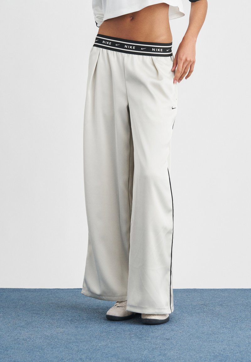 Λευκό παντελόνι φόρμας Nike με μαύρη μέση με λογότυπο, φαρδιά πόδια και ρίγες στο πλάι. Το ύφασμα φαίνεται λείο με ελαφριά γυαλάδα.