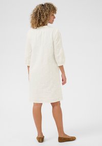 Vit texturerad klänning med blommönsterbroderi, trekvarts puffärmar och en l�ätt utställd fåll. Kombinerad med bruna platta skor.