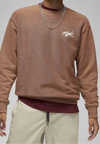 Sweat-shirt marron texturé avec poignets et ourlet inférieur côtelés, arborant un logo de marque blanc sur la poitrine, superposé à une chemise bordeaux.