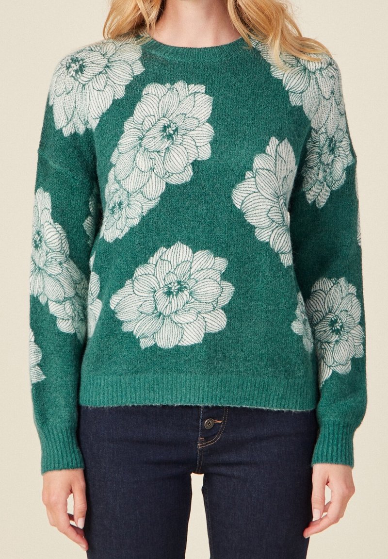 Pull vert tricoté avec de grands motifs floraux blancs porté avec un jean bleu foncé, montré des épaules aux hanches.