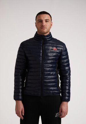 Marineblå puffjakke med høj krave, quiltet design, sidelommer og et logo patch på brystet. Glat, skinnende tekstur. Sorte detaljer.