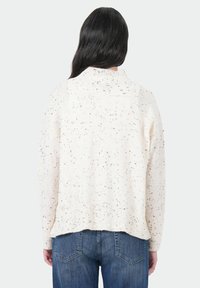 Crèmekleurige gebreide trui met gesprenkelde zwarte accenten, geribbelde onderkant en hoge kraag, gecombineerd met een blauwe denim jeans. De trui heeft een relaxte pasvorm.