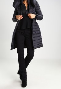 Giacca a vento nera imbottita con colletto alto e chiusura con cerniera, abbinata a una maglia e pantaloni neri. Il tessuto è liscio, con un motivo trapuntato.