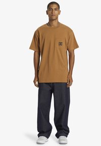 Muž ve hnědém oversized tričku s kapsou, volných tmavých džínách, bílých teniskách a stříbrných špercích, stojící s neutrálním výrazem.