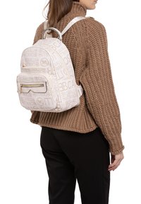 Zaino bianco con motivo a logo ripetuto, dotato di tasca frontale con zip e spallacci regolabili, indossato sopra un maglione in maglia marrone.