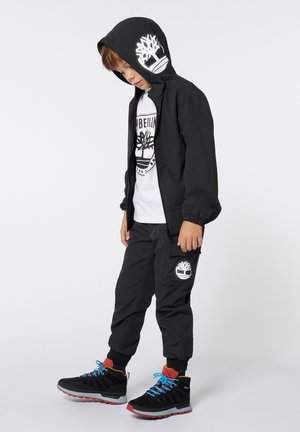Veste à capuche noire avec des poignets élastiques, logo blanc ; t-shirt blanc en dessous ; pantalon cargo noir avec logo ; bottes noires avec lacets bleus.