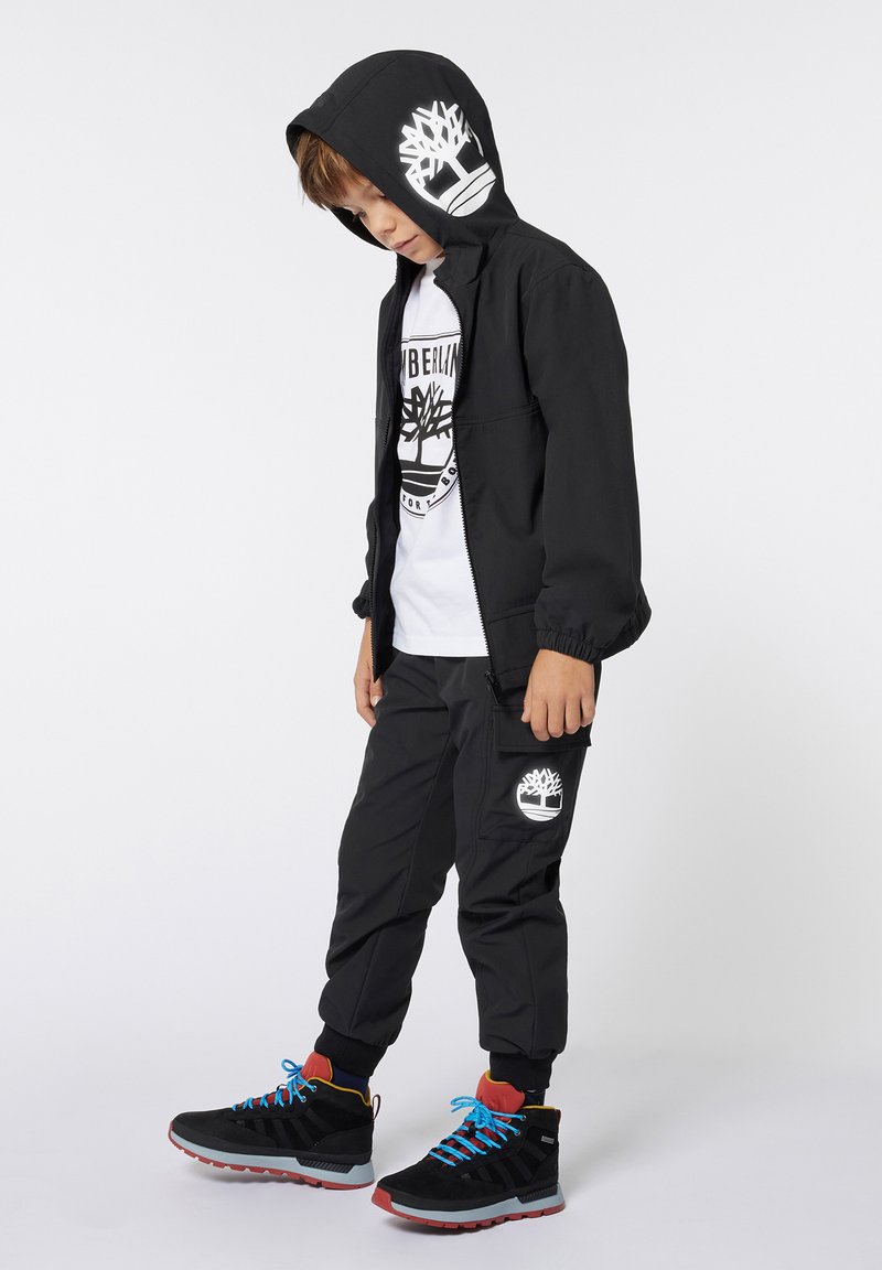 Veste à capuche noire avec des poignets élastiques, logo blanc ; t-shirt blanc en dessous ; pantalon cargo noir avec logo ; bottes noires avec lacets bleus.