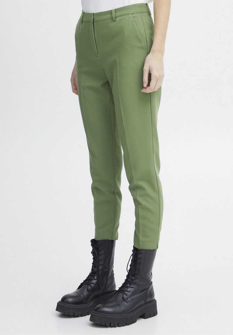 Personne portant un pantalon slim vert olive et des bottes de combat noires lacées, debout devant un fond clair uni.