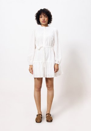 Robe en coton blanc à manches longues, avec une fermeture à boutons sur le devant et une taille nouée. Présente un ourlet volanté et des détails brodés subtils. Sandales brunes.