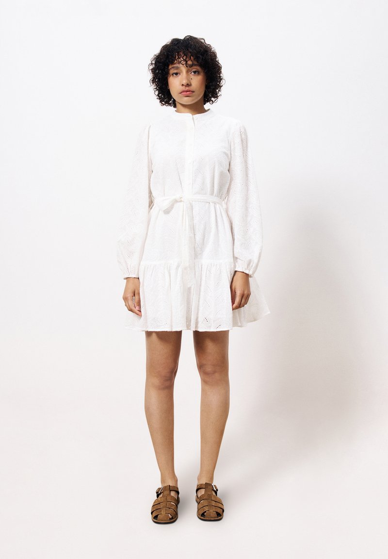 Robe en coton blanc à manches longues, avec une fermeture à boutons sur le devant et une taille nouée. Présente un ourlet volanté et des détails brodés subtils. Sandales brunes.