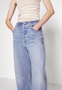 Jasnoniebieskie dżinsowe spodnie o szerokich nogawkach z wybledzonym wykończeniem, wysoki stan, kieszenie z przodu i subtelne przeszycia. Nosi się z beżową koszulą bez rękawów w paski.