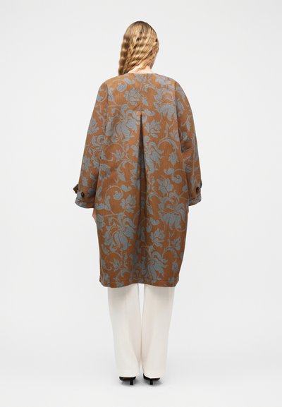 Γυναίκα με μακριά κυματιστά μαλλιά, φορώντας παλτό με καφέ και γκρι λουλουδάτο μοτίβο, λευκό παντελόνι και μαύρες γόβες, γυρισμένη προς τα πίσω σε λευκό φόντο.