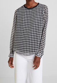 Blusa a scacchi bianco e nero con maniche lunghe trasparenti, scollatura rotonda e polsini neri, abbinata a pantaloni bianchi a gamba larga.
