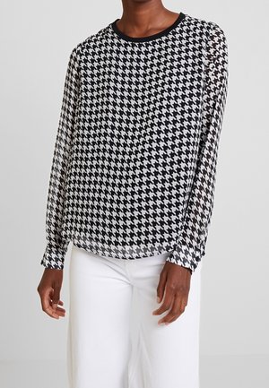Blusa a scacchi bianco e nero con maniche lunghe trasparenti, scollatura rotonda e polsini neri, abbinata a pantaloni bianchi a gamba larga.