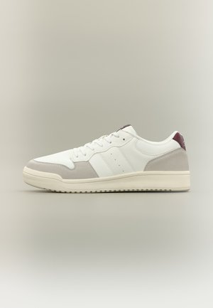 Pantof sport alb, tip low-top, cu talpă crem, accente din piele întoarsă gri, șireturi albe și o clapetă roșu închis în zona călcâiului, pe un fundal neutru.