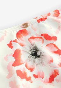 Biela látka s veľkým kvetinovým vzorom v odtieňoch ružovej a červenej. Blízko vrchu sa nachádza malý zlatý logo detail.