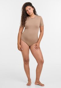 Beige Bodysuit mit kurzen Ärmeln, körperbetonter Passform, weicher Textur und rundem Ausschnitt. Das Model steht barfuß vor einem schlichten Hintergrund.
