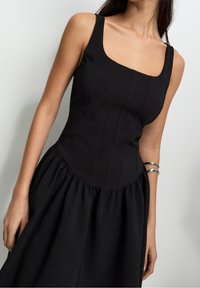 Robe noire avec un corsage ajusté, un décolleté carré et une jupe évasée et froncée. Tissu lisse avec des coutures verticales et une texture fluide.