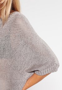 Personne blonde portant un pull en tricot léger gris clair, ample, avec des sous-vêtements noirs visibles en dessous, sur un fond uni.