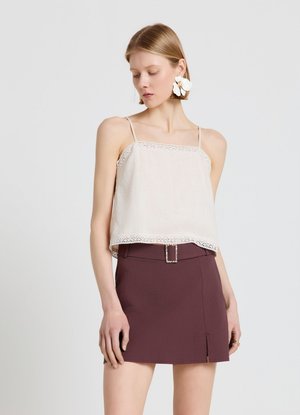 Jeune femme portant un haut blanc sans manches avec bordure en dentelle, une minijupe violet foncé avec ceinture, et de grandes boucles d'oreilles florales blanches.