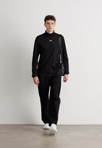 Svart langermet skjorte med høy krage og logo, kombinert med svarte cargo-bukser. Skoene er hvite sneakers. Enkelt, moderne design.