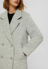 Woman wearing a light gray textured double-breasted coat with lapels, buttons, and side pockets, hand tucked in pocket, black top underneath.