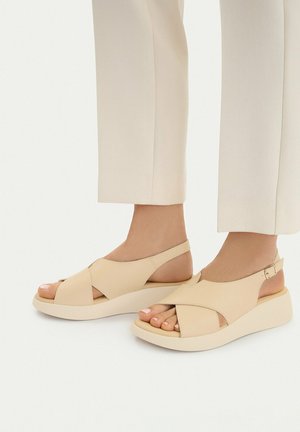 Stopy w beżowych sandałach typu slingback z odkrytymi palcami, szerokimi krzyżującymi się paskami i grubymi podeszwami, zestawione z jasnymi, krótkimi spodniami.