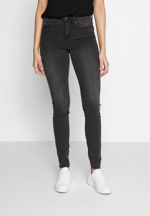 Person iført sorte skinny jeans, sort top og hvide sneakers, stående mod en ensfarvet hvid og grå baggrund.