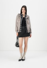 Mlada žena nosi sivi hoodie preko crno-kariranog košulje i crne suknje, drži crnu torbicu i nosi bež čarape sa crnim potpeticama.