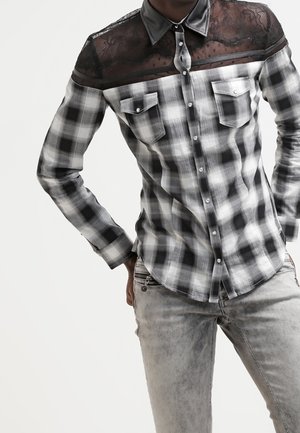 Man draagt een zwart-wit-grijs geruit overhemd met kant bij het schouderstuk en een leren kraag, gecombineerd met lichtgrijze jeans.