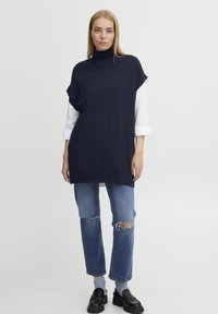 Maglione a collo alto oversized blu navy con maniche corte, indossato sopra una camicia bianca, abbinato a jeans blu strappati e mocassini neri.