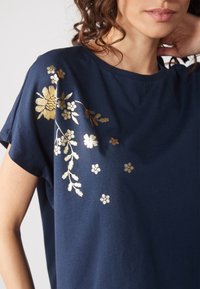 Femme portant une chemise bleu marine avec un motif floral doré sur l'épaule et le haut de la poitrine, main reposant près du cou, cheveux bouclés visibles.