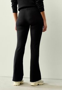 Schwarze, ausgestellte Leggings aus dehnbarem Material, mit glatter Oberfläche und hohem Bund, kombiniert mit hellen Sportschuhen.