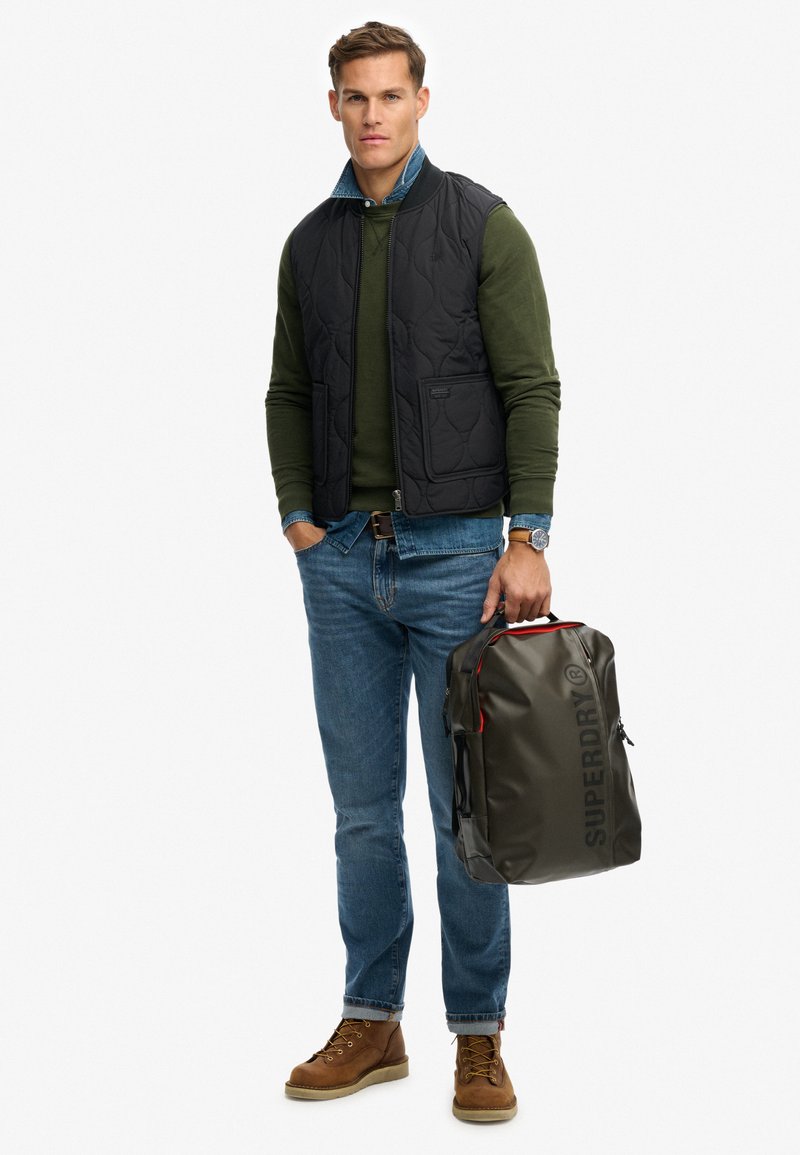 Pikowana czarna kamizelka, zielony sweter, niebieskie dżinsy i brązowe buty; trzymając ciemnozielony plecak z czerwonymi akcentami i napisem "SUPERDRY".