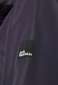 Tessuto impermeabile blu navy con un patch nero testurizzato a forma di quadrato che presenta il testo bianco "Jack Wolfskin". La superficie appare liscia e lucida.