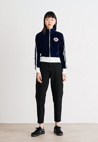 Giacca da track in velluto blu navy con dettagli bianchi e logo, abbinata a pantaloni neri a sigaretta e sneakers nere. Design semplice e sportivo.