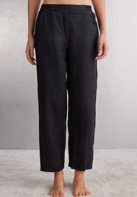 Zwarte linnen broek met een elastische tailleband, losse pasvorm, rechte pijpen en een zachte textuur, gepresenteerd op een model zonder schoenen.