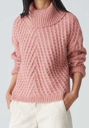 Femme portant un pull à col roulé rose torsadé avec manches retroussées et pantalon blanc, mains dans les poches.