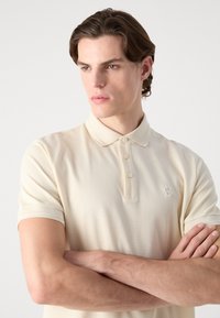 Polo skjorte i lys beige, korte ermer, laget av teksturert stoff. Har en placket med tre knapper og en liten brodert logo på brystet.