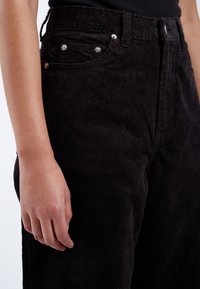 Pantalones de pana negros con una cintura alta, textura de rayas verticales y herrajes en plata. Incluyen bolsillos frontales y una forma ajustada.