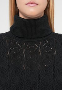 Juodas megztas golfas su tekstūriniais raštais, apimančiais rombo ir lapų motyvus, pabrėžiantis ribotą apykaklę ir minkštą audinį.