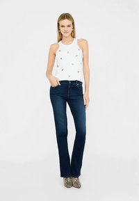 Bílý tílko bez rukávů s vyšívanými ozdobami, doplněné tmavě modrými rozšířenými džínami a vzorovanou obuví. Hladká látka, přiléhavý střih.